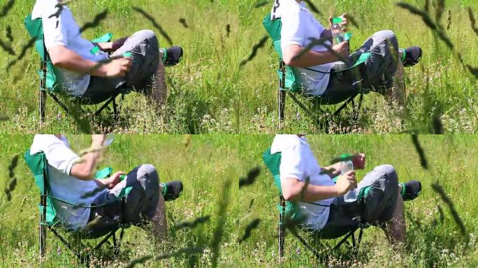 一名男子坐在折叠野餐椅上，用智能手机工作。他休息一下，喝瓶子里的水。周围是被风倾斜的草。