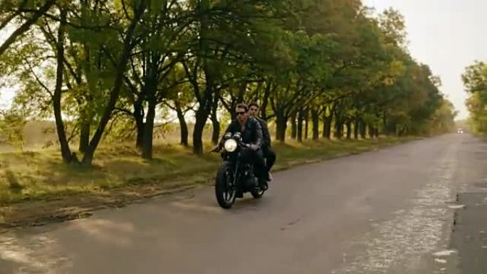 秋天在森林路上骑摩托车的年轻酷夫妇的跟踪镜头。戴着墨镜的迷人年轻人开着直升机，而他的女朋友坐在后面