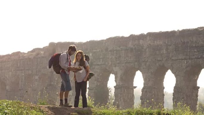 年轻可爱的夫妇背包客游客阅读地图指向罗马渡槽拱门在罗马公园废墟上浪漫的迷雾日出，吉他和睡袋慢动作