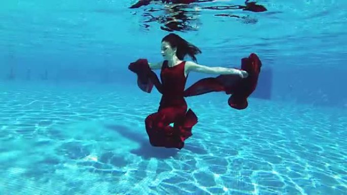 一个红头发的美丽新娘女孩在阳光明媚的日子里，穿着蓝色背景的红色连衣裙在室外游泳池里游泳和摆姿势。
