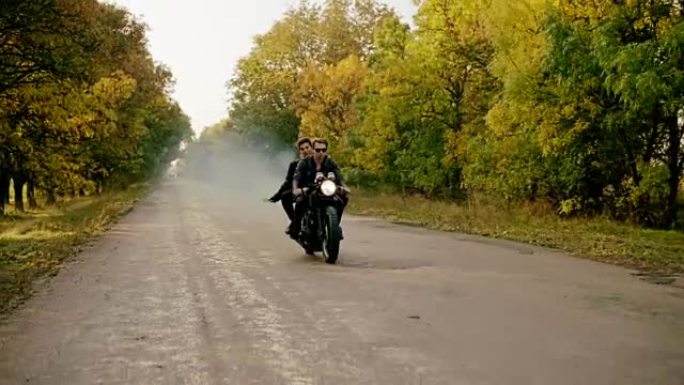 秋天，戴着墨镜的帅哥和女友骑着摩托车在森林的柏油路上。他的女朋友拿着一枚烟雾弹。慢动作