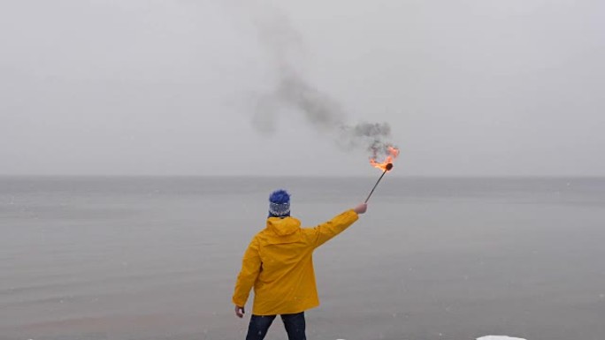 在冰冻的大海背景下，穿着黄色外套的火把的人的后视图。雪落在前景