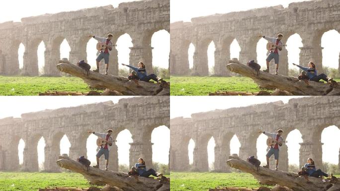 幸福的年轻夫妇背包客游客在原木树干上，用手在罗马帕尔科·德格里·阿奎多蒂公园的古罗马渡槽废墟前指向方