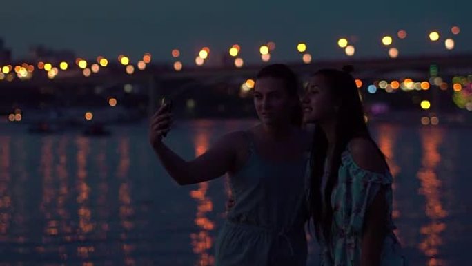 两个女朋友晚上在灯光的背景下在河边自拍。慢动作