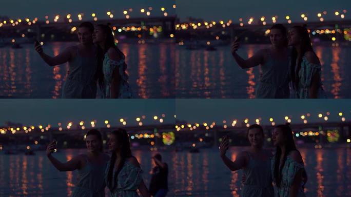 两个女朋友晚上在灯光的背景下在河边自拍。慢动作
