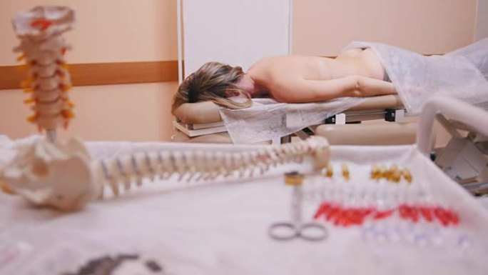 脊椎按摩师按摩一名躺在按摩台上的年轻女子，推动前景中的腰椎、脊柱模型和药物