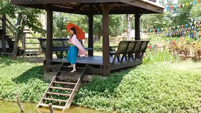 亚洲妇女穿着泰国传统文化服装，复古风格的红色雨伞走到河边的凉亭，坐下来敬礼 (sawasdee)。