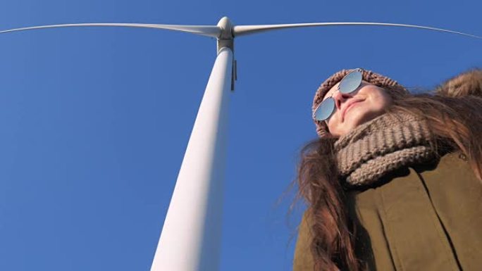 戴着太阳镜的年轻女性站在风车的大叶片下，对着天空