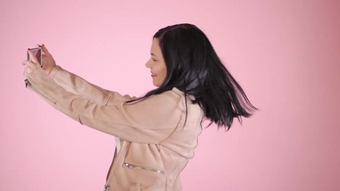 微笑快乐的黑发女人，穿着粉色夹克，在彩色背景下用智能手机自拍。慢动作