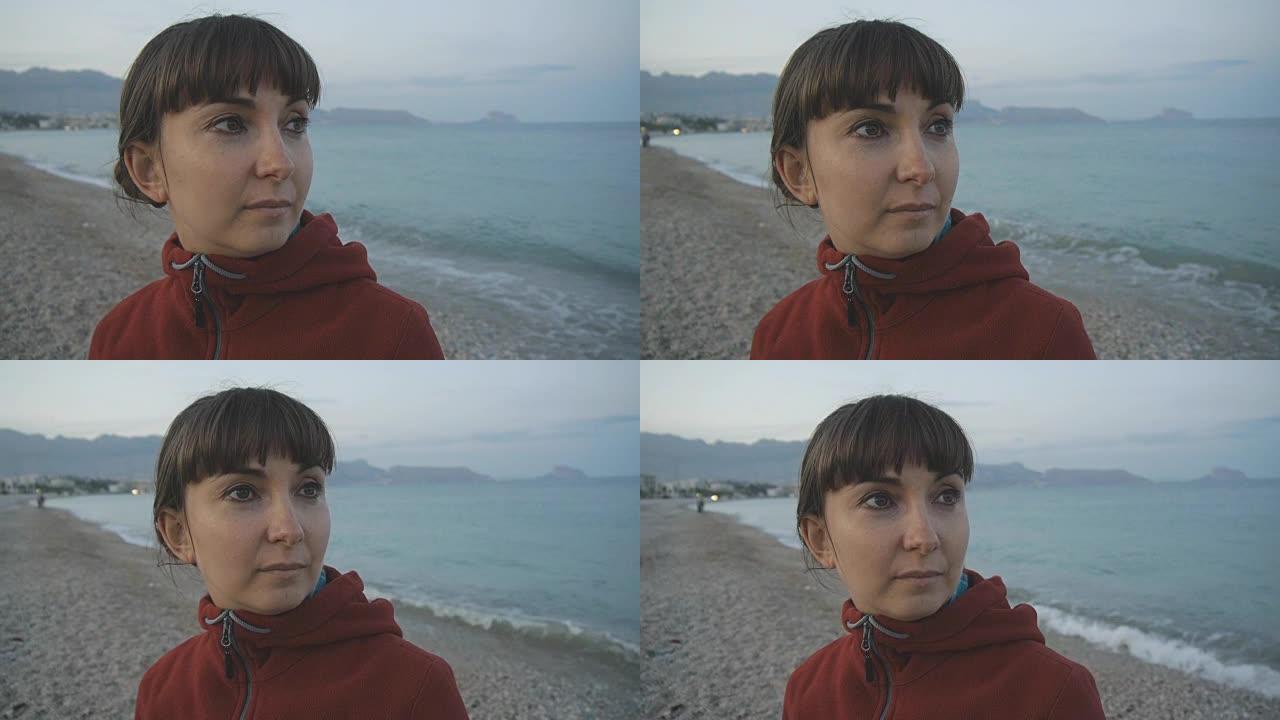 空荡荡的海滩背景上的年轻女子。迷人的高加索棕色头发女性的特写镜头，穿着红色连帽衫，看着大海。