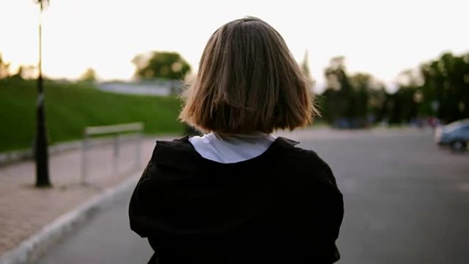 背面跟踪女孩在空荡荡的街道上行走的镜头。戴上黑色耳机，回头看她的肩膀。对着镜头微笑。快乐，快乐。夏。