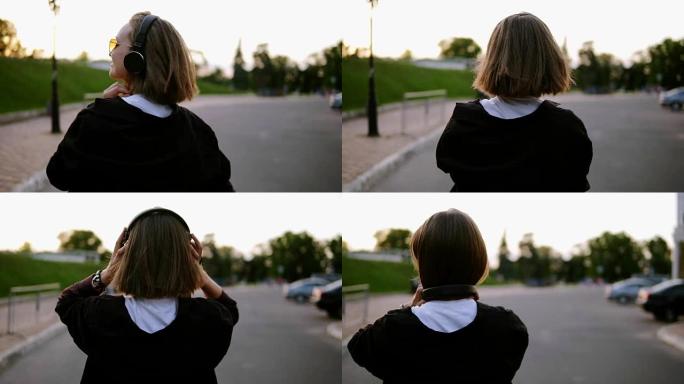 背面跟踪女孩在空荡荡的街道上行走的镜头。戴上黑色耳机，回头看她的肩膀。对着镜头微笑。快乐，快乐。夏。