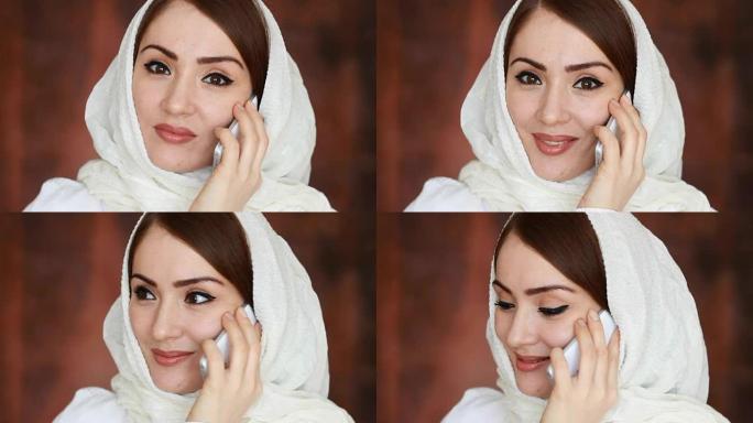 一个美丽的阿拉伯女人在传统的头巾头巾的肖像特写在手机上交流。一名穆斯林女孩在白色智能手机上讲话并微笑