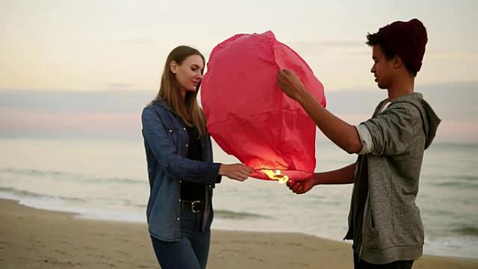 年轻的多民族夫妇在发射前手持红色纸灯笼。海滩上的浪漫约会。有魅力的女人和她的非洲男友一起拿着火灯放飞