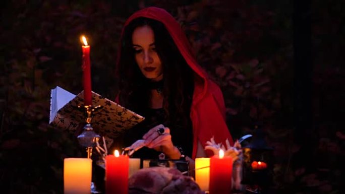 迷人的美丽黑发女巫穿着红色斗篷在神秘的秋天森林中让人联想到。带有心脏，鸡腿和豪猪针的巫术