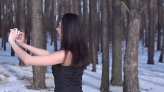 穿着黑色连衣裙的年轻黑发女孩在冬天的森林里用智能手机自拍，微笑着转身