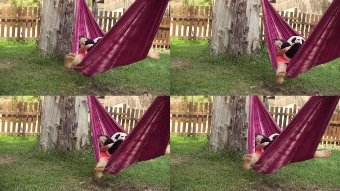 夏令营中年轻快乐的小女孩躺在五颜六色的吊床上放松在外面玩绿草地上的plushes狗娃娃儿童慢动作