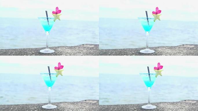 在马提尼酒杯中搭配蓝色利口酒的鸡尾酒，配以两根黑色吸管，并装饰着星光和粉红色的花瓣，背景为大海