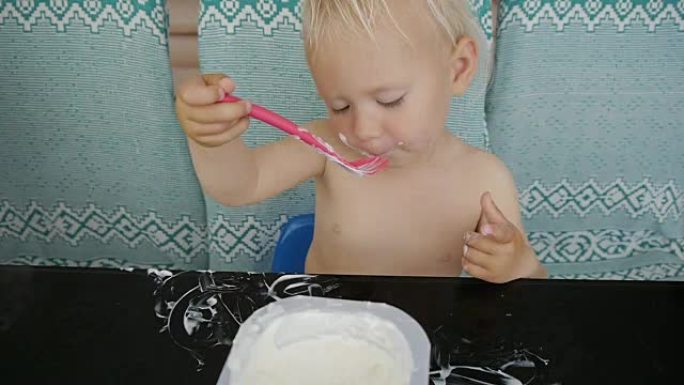 宝宝吃酸奶弄得一团糟。可爱的白人小女孩坐在黑色玻璃桌子旁舔着勺子里的酸奶。