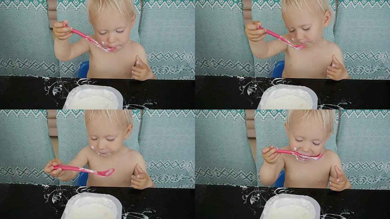 宝宝吃酸奶弄得一团糟。可爱的白人小女孩坐在黑色玻璃桌子旁舔着勺子里的酸奶。
