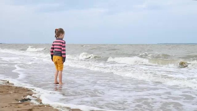 美丽的小女孩看着大海上的海浪站在水面附近。海上弱风暴