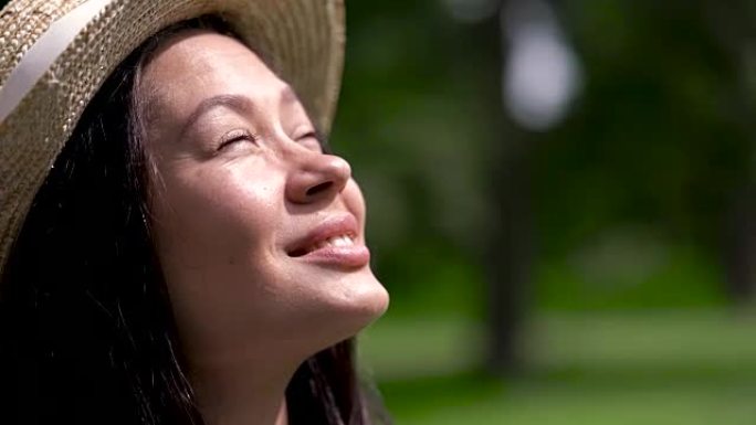 一个戴着夏日帽子的微笑的亚洲女孩的特写镜头，看着太阳，眨着眼睛。