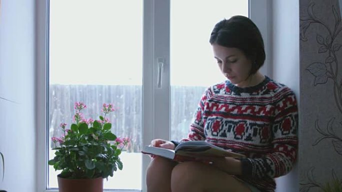美丽的年轻黑发女人穿着针织毛衣看书，坐在窗边。背景中模糊的飘落的雪。