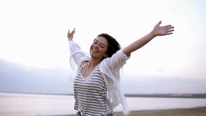 一个美丽的卷发黑发女人的肖像，伸出双手快乐地在海边或湖边奔跑。穿着白色休闲服。微笑，快乐，自由