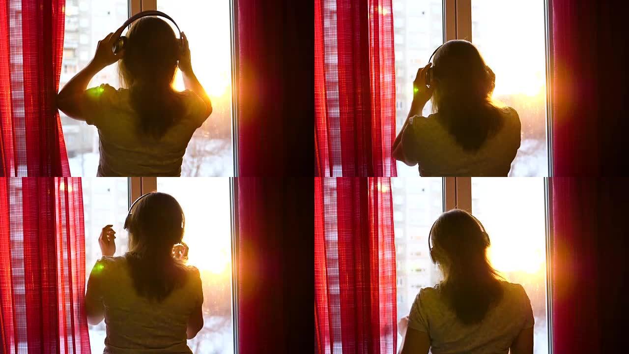 一个年轻女孩来到窗前，戴着耳机听音乐。日落背景模糊，女人在耳机中欣赏旋律。太阳的光线穿过玻璃
