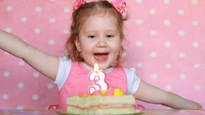 一个可爱的小女孩许愿，在聚会上吹灭生日蛋糕上的蜡烛。可爱的孩子。儿童假期的概念。3年，粉色背景。