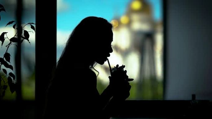 年轻女子的黑色剪影在咖啡馆里用稻草从玻璃上喝鸡尾酒，靠窗
