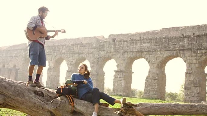 幸福的年轻夫妇背包客游客在罗马浪漫公园的古罗马渡槽废墟前弹吉他唱歌日出慢动作