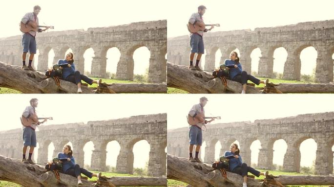 幸福的年轻夫妇背包客游客在罗马浪漫公园的古罗马渡槽废墟前弹吉他唱歌日出慢动作