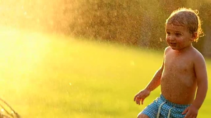 小男孩享受着120帧慢镜头在日落黄金时间里被溅起的水花。蹒跚学步的婴儿走向洒水车，享受生活中的小事情