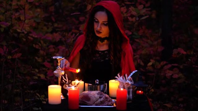迷人的美丽黑发女巫，穿着红色斗篷的书，在神秘的秋天森林中让人联想到。带有心脏，鸡腿和豪猪针的巫术