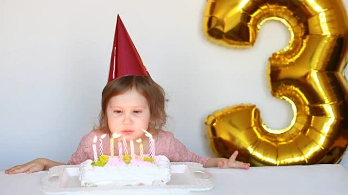 有趣的快乐孩子许愿，在派对上吹灭生日蛋糕上的蜡烛。戴着粉红色帽子的可爱的小女孩微笑着，对假期感到满意
