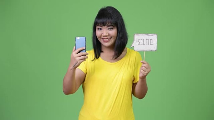 美丽快乐的亚洲女人拿着手机和自拍纸标志
