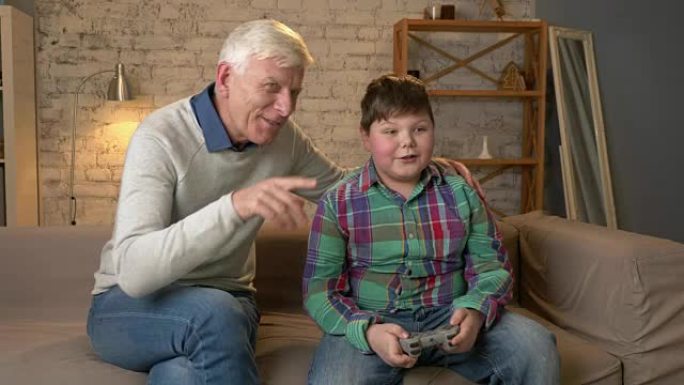 爷爷在游戏机游戏中享受孙子的胜利。老人和一个年轻的胖男孩坐在沙发上。电子游戏。家庭舒适，家庭田园诗，
