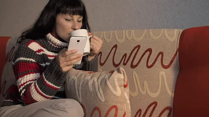 穿着脱衣毛衣的年轻女子坐在沙发上，使用智能手机，喝咖啡。女孩正在聊天，写博客，查看电子邮件。在线营销