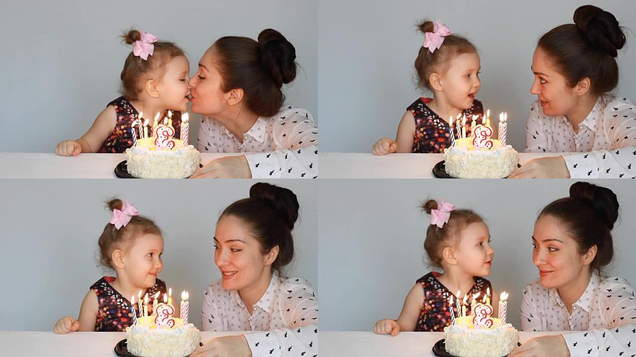 快乐的妈妈和孩子在聚会上看着生日蛋糕上蜡烛的火焰。母亲祝贺、拥抱和亲吻她的女儿。3年