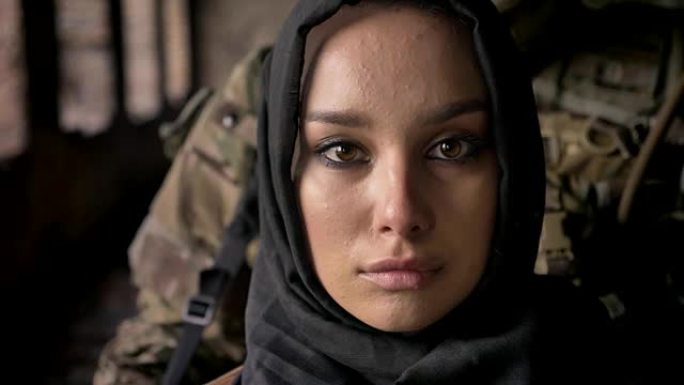 近距离拍摄的年轻穆斯林妇女戴着头巾看着镜头，武装士兵与枪站在妇女身后，军事概念