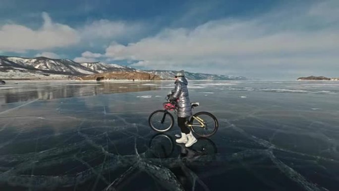 女人正走在冰上的自行车旁边。这个女孩穿着银色羽绒服，背包和头盔。冰冻的贝加尔湖的冰。自行车上的轮胎上