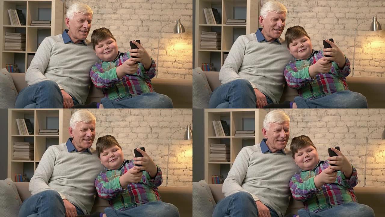 祖父和孙子坐在沙发上使用智能手机，进行自拍。年轻的胖孩子和祖父微笑着。家居舒适，家庭田园诗，舒适概念