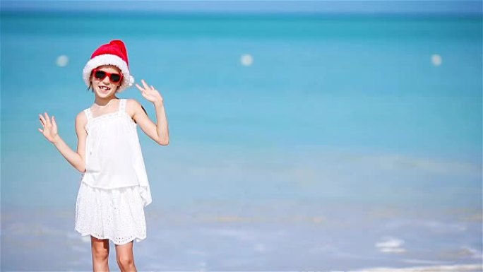 圣诞节假期，白色海滩上戴着圣诞帽的可爱小女孩。快乐微笑的孩子看着相机