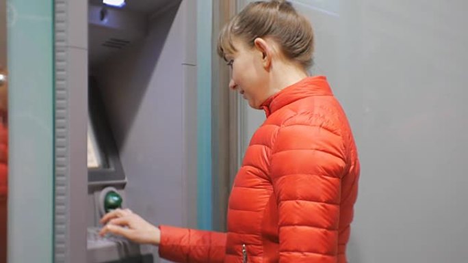 使用提款机的女人。穿着红色泡泡夹克的迷人年轻女性将信用卡插入自动取款机。微笑着输入密码的女人。