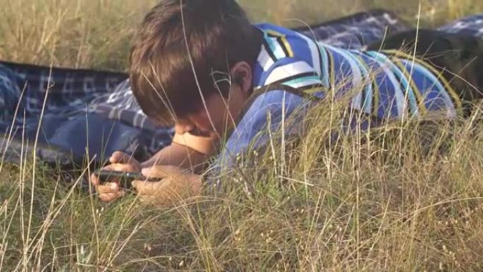 小男孩看智能手机男孩晚上躺在草地上的地毯上，挥动双腿