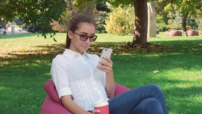 休闲年轻女子坐在城市公园喝咖啡，使用智能手机