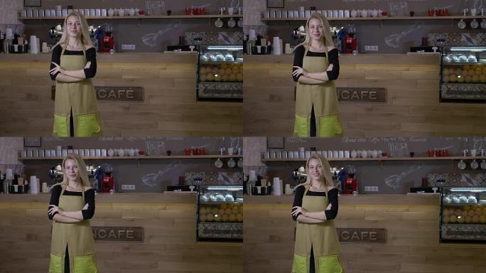 努力工作的年轻金发女子终于开办了自己的咖啡馆生意，自豪地站在柜台前，双臂交叉，微笑着