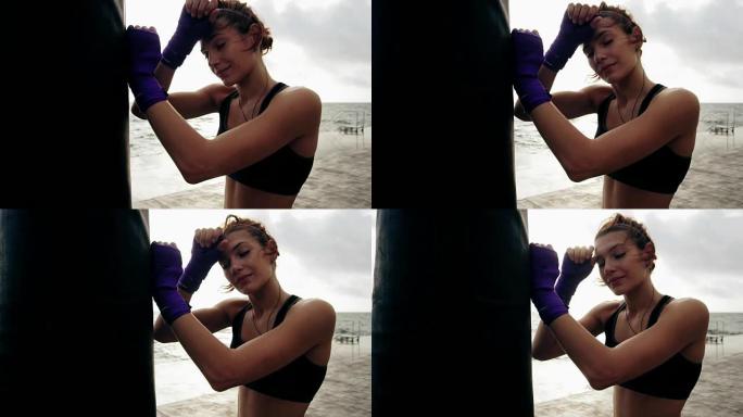 特写镜头，一名年轻女子在拳击袋对着太阳进行艰苦训练后休息。她的手被紫色的拳击胶带包裹着。夏天在海滩上