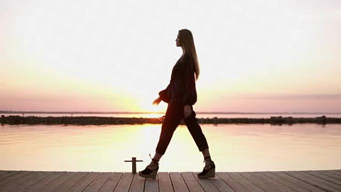 慢镜头，镜头从一边美丽的长头发的黑衣女子走在木码头。清晨黄昏，阳光洒在后台